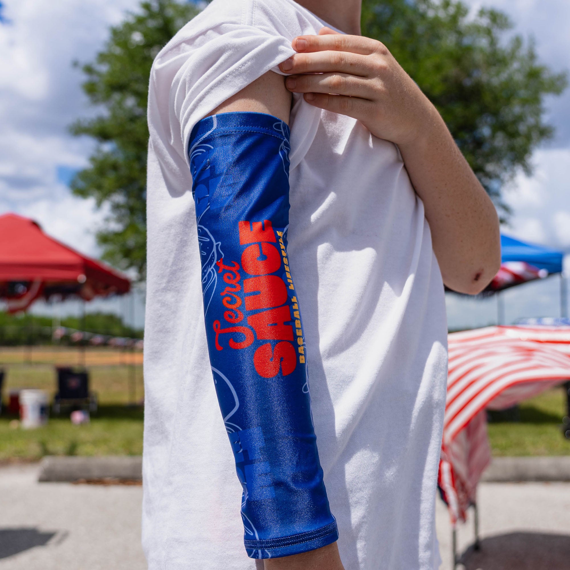 Close-up of youth arm showing blue Secret Sauce arm sleeve with full Secret Sauce Baseball Lifestyle logo visible while pulling up sleeve over white tee at outdoor event