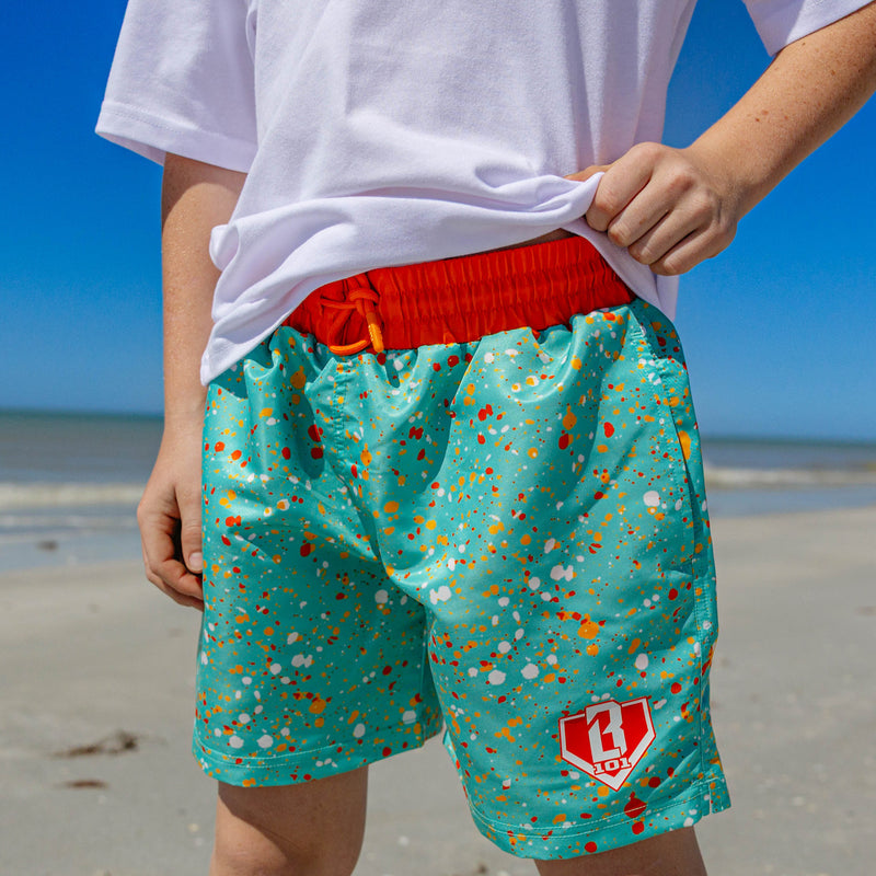 Close-up of BL101 teal splatter print swim trunks lifted to show the orange built-in compression liner and BL101 logo on the front pocket.
