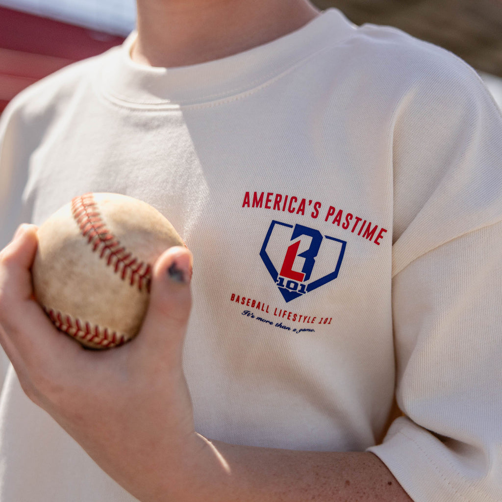  Close-up of the BL101 America's Pastime chest logo on the youth cream tee, with a baseball held in the foreground.