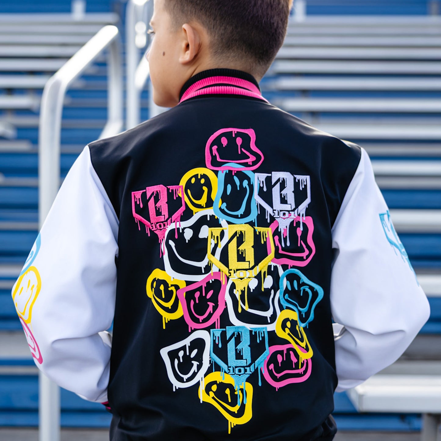 Person wearing a black varsity jacket with colorful smiley faces, and yellow BL101 logo standing in front of blue bleachers.