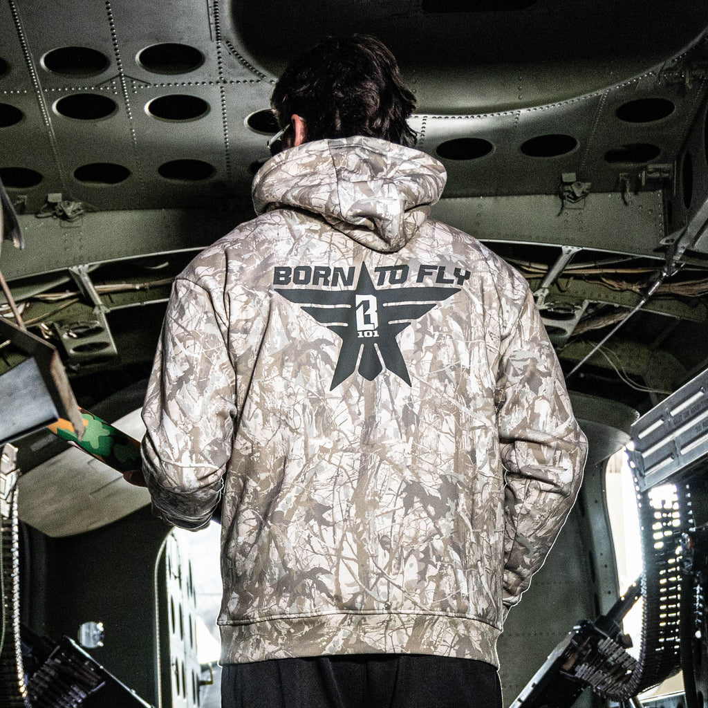 Person wearing a camouflage hoodie with 'Born to Fly' text inside an aircraft.