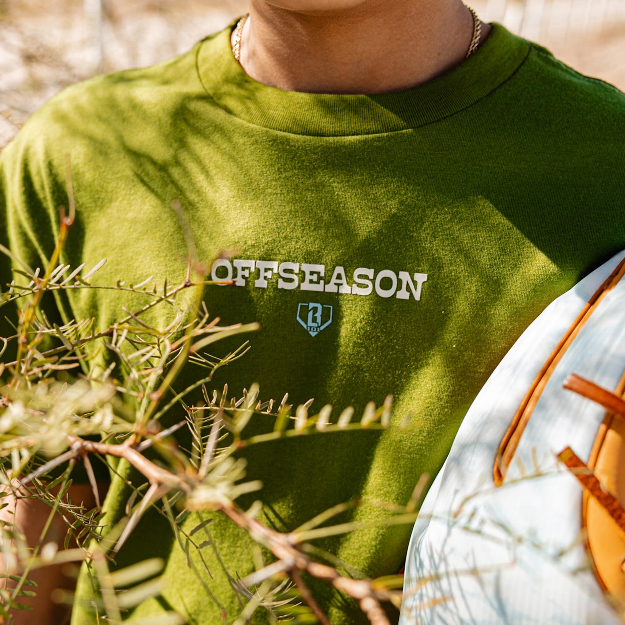 Close up of person wearing a green 'OFFSEASON' t-shirt with a BL101 logo, surrounded by nature.