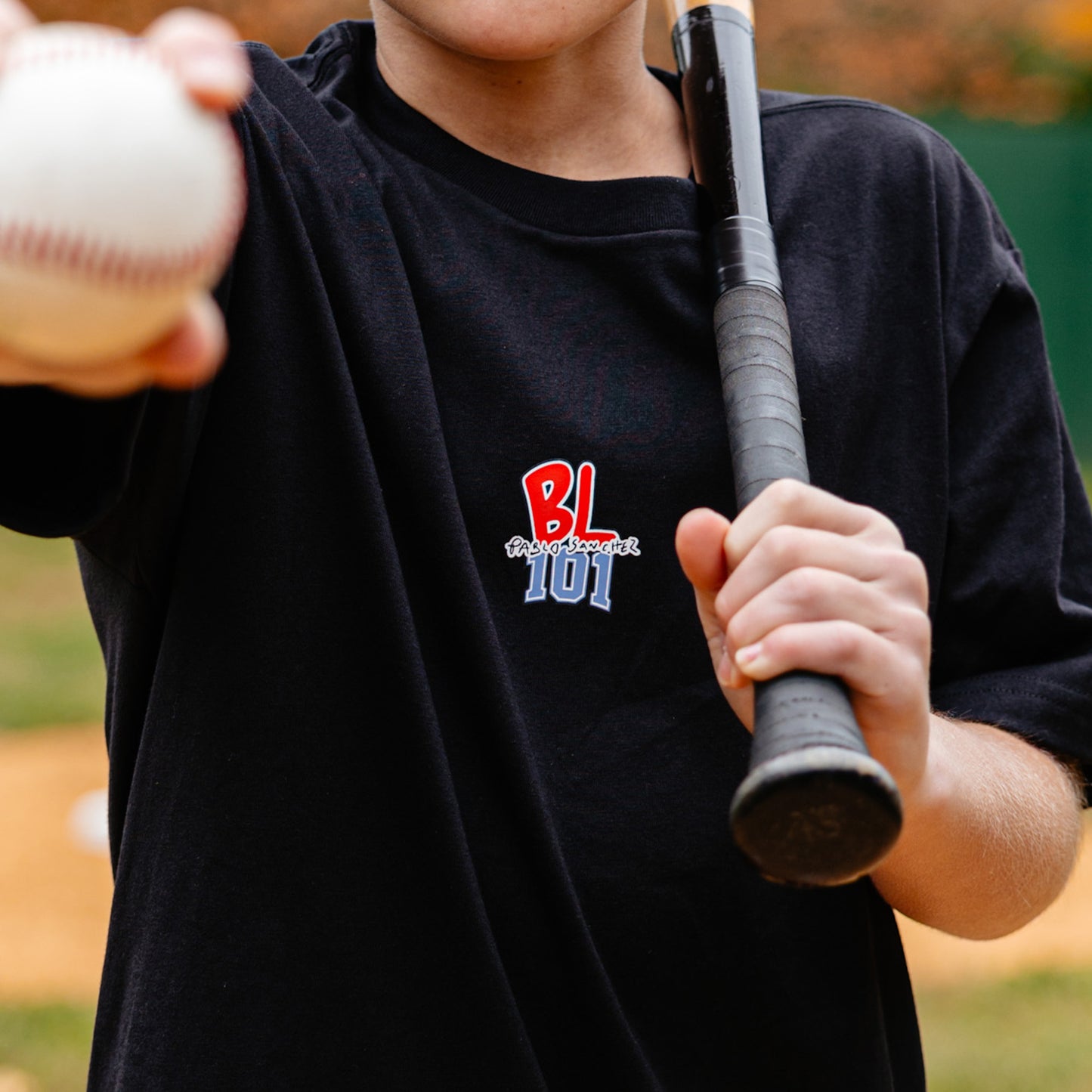 Person holding a baseball and bat with a black shirt featuring a BL101 Pablo Sanchez logo.