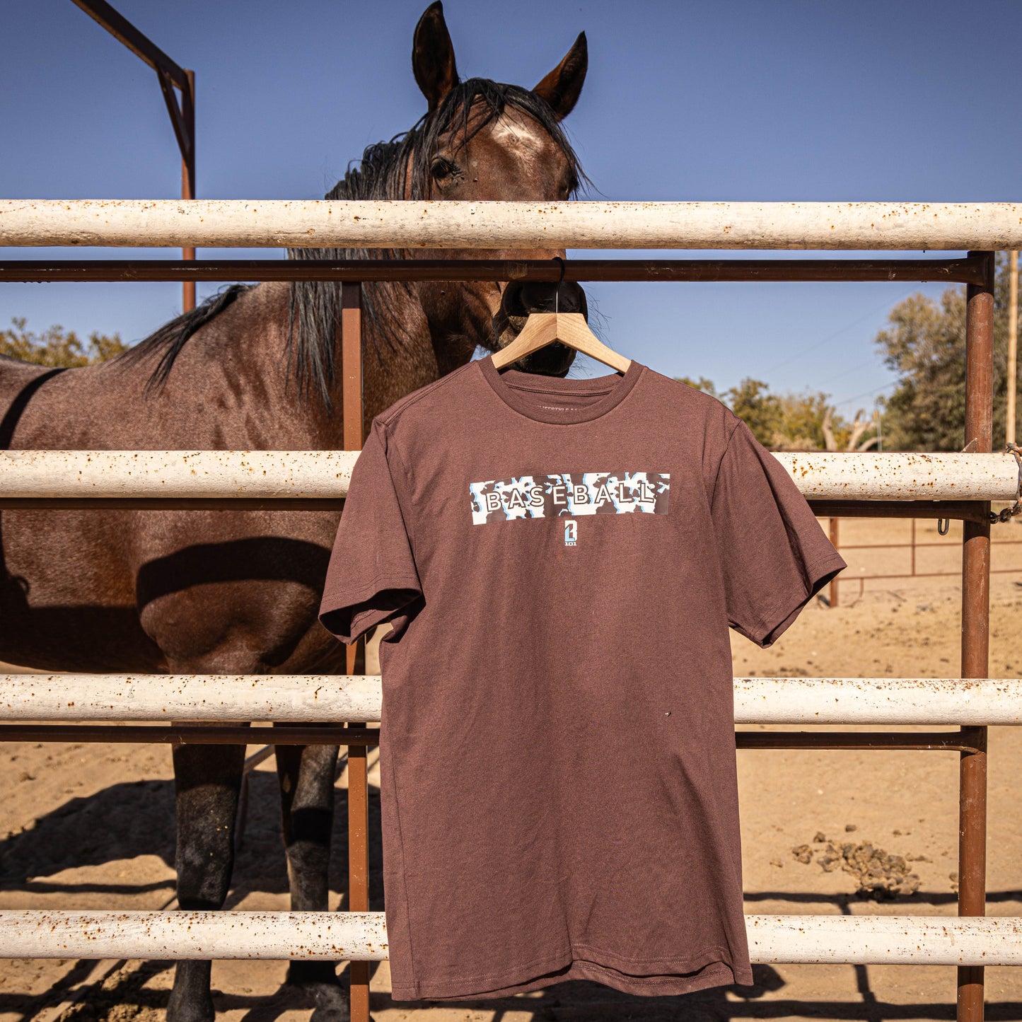 Brown t-shirt with cow graphic design hanging on a wooden post with a horse in the background