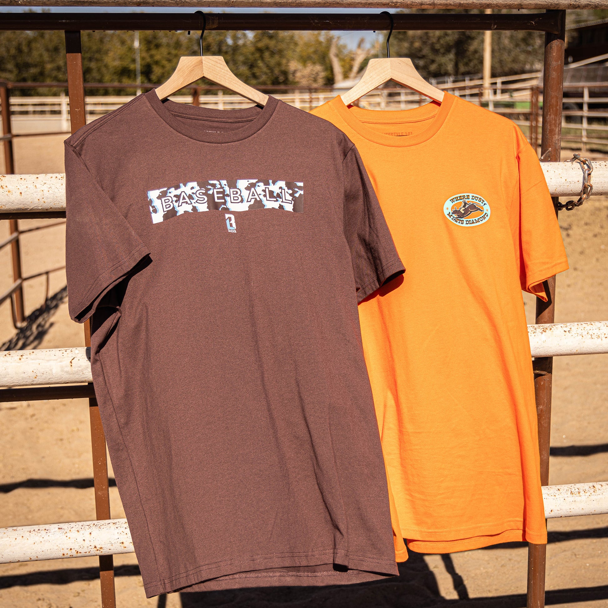 Two t-shirts, one brown and one orange, hanging on a wooden rack with a fence and trees in the background.