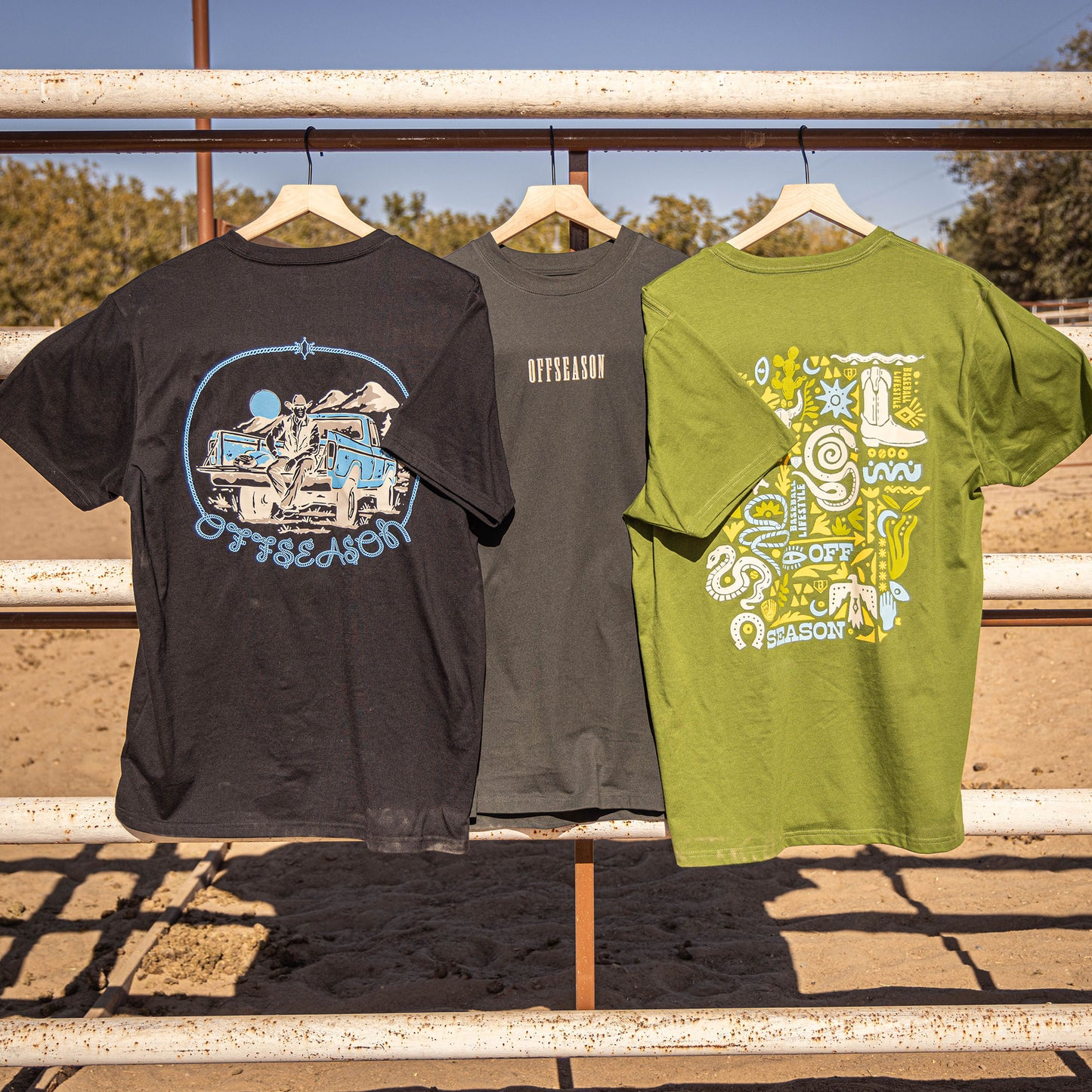 Three black, grey and green t-shirts with different desert designs hanging on a metal railing outdoors.