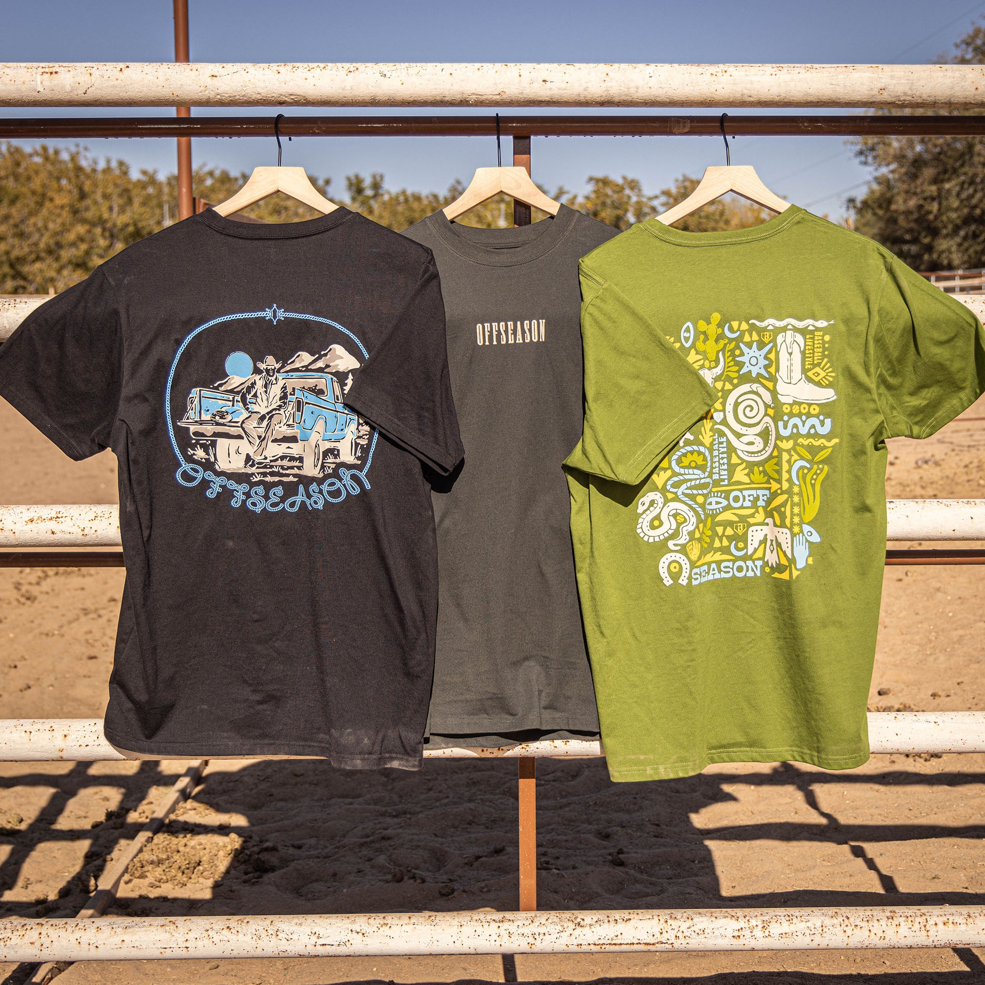 Three black, grey and green t-shirts with different desert designs hanging on a metal railing outdoors.