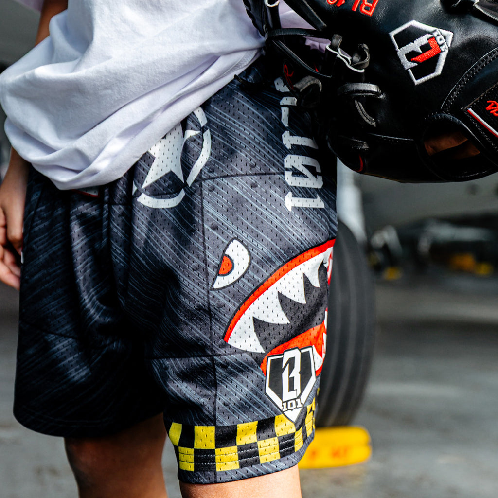 Close up of Military-inspired black athletic shorts featuring shark mouth graphics, a white star emblem, a red ‘Keep Clear’ detail, white BL101 logos on the sides, a green waistband, and yellow checkerboard trim.