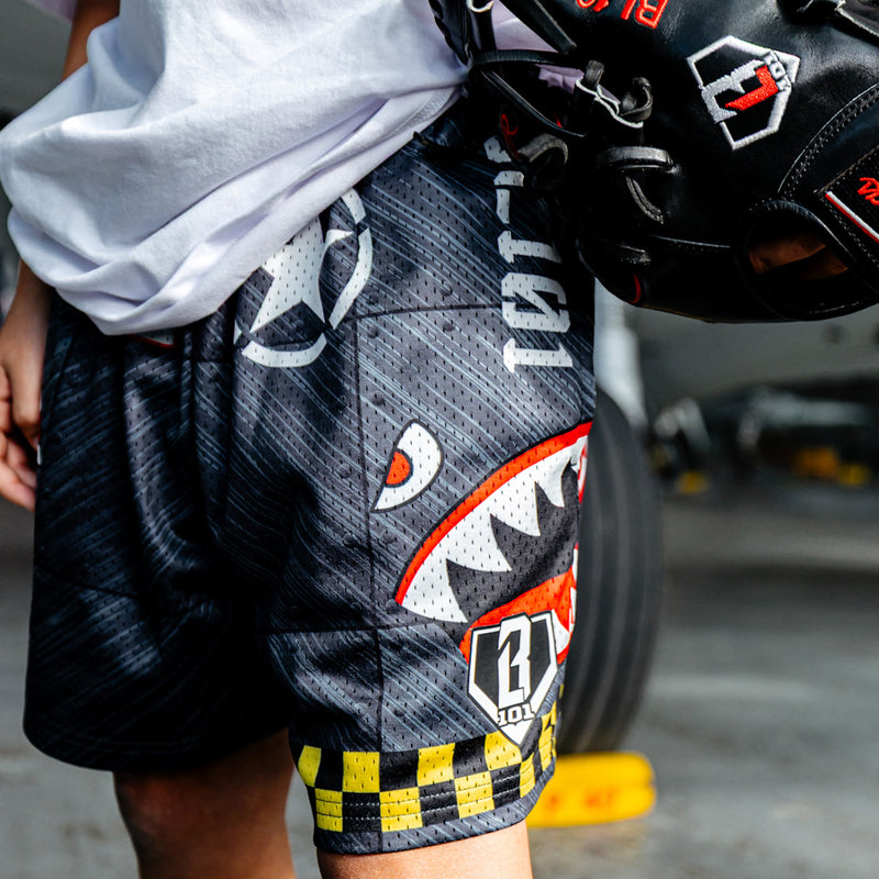 Close up of Military-inspired black athletic shorts featuring shark mouth graphics, a white star emblem, a red ‘Keep Clear’ detail, white BL101 logos on the sides, a green waistband, and yellow checkerboard trim.