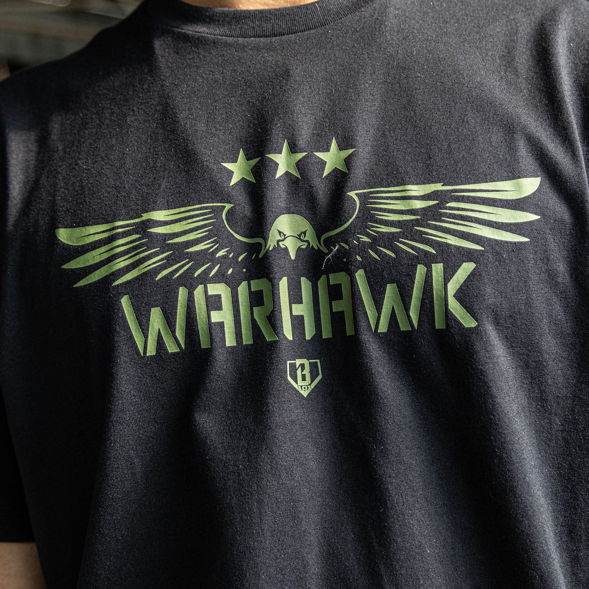 Close up of Youth black t-shirt featuring an army green eagle graphic with green “WARHAWK” lettering and a green Armor BL101 logo