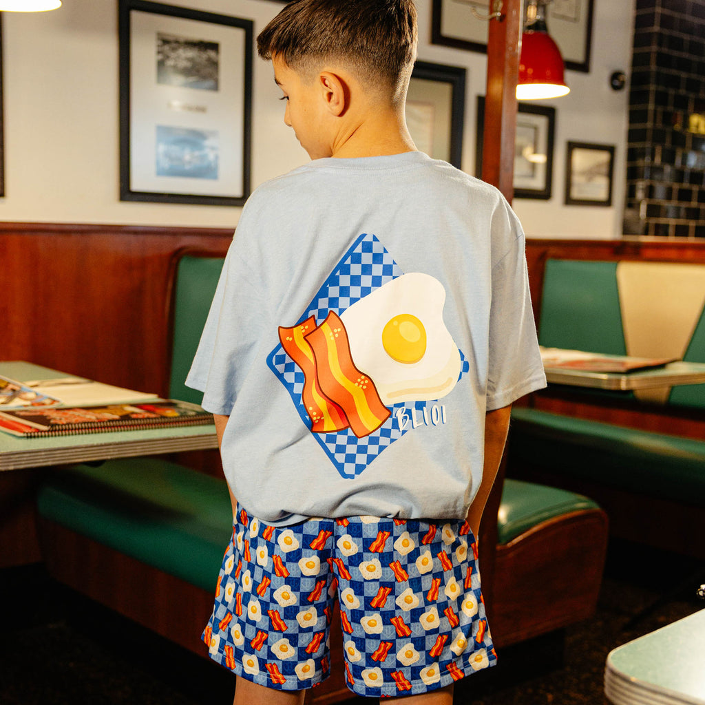 Child wearing a blue shirt and blue checkered shorts with bacon and egg design in a diner setting