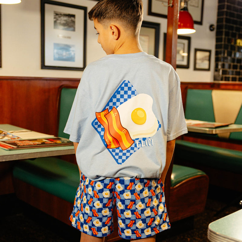 Child wearing a blue shirt and blue checkered shorts with bacon and egg design in a diner setting