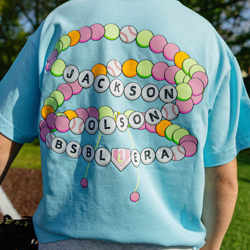 Back view of the youth light blue BSBL Era tee showing the colorful friendship bracelet graphic with "Jackson Olson BSBL Era" lettering and baseball bead details