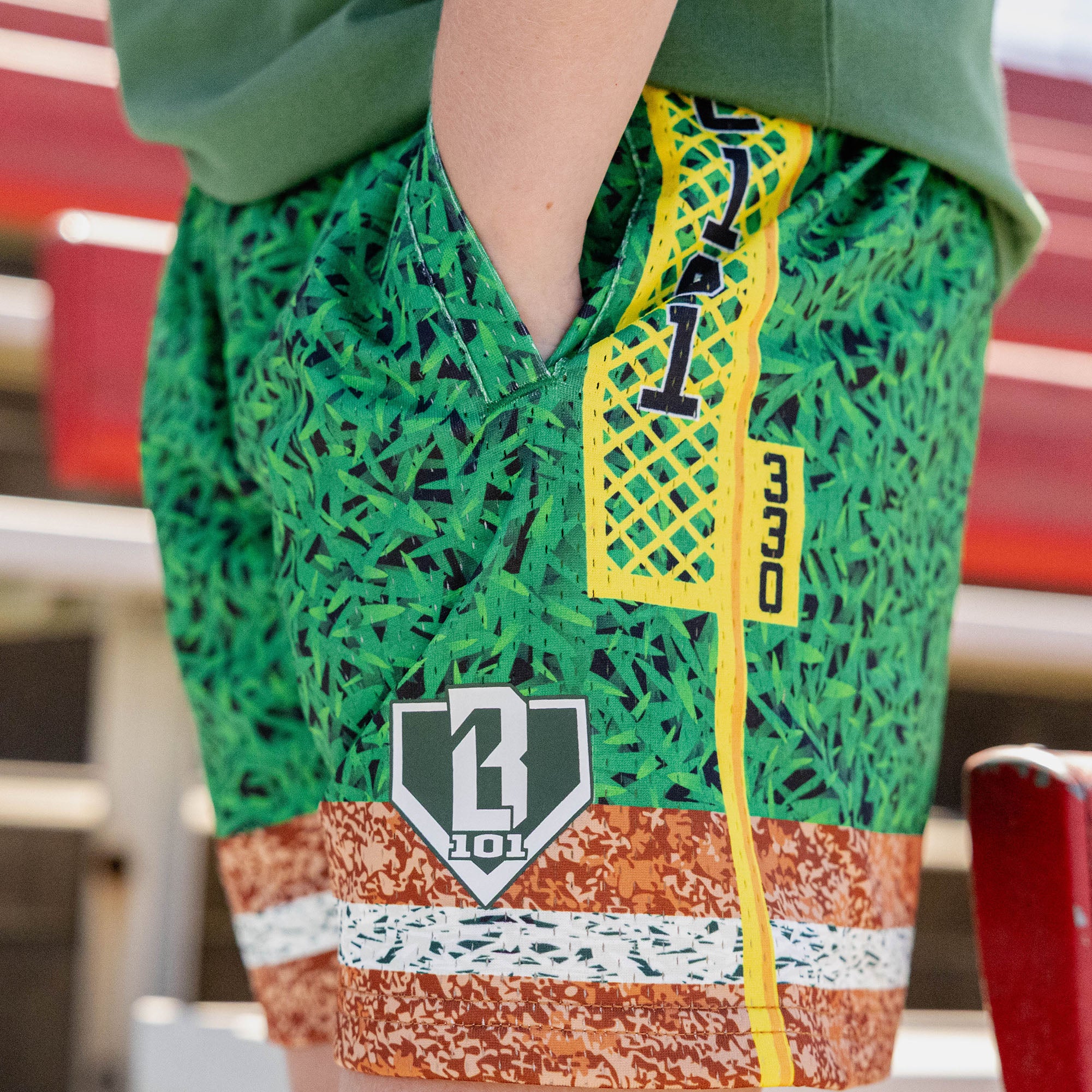 Close up of green grass print mesh shorts with yellow foul pole side stripe BL101 logo patch and brown marbled hem