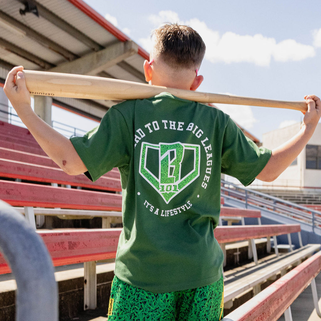 Back view of boy holding a bat across his shoulders at red stadium bleachers wearing a forest green tee with Backyard to the Big Leagues graphic featuring a grass textured home plate BL101 logo and It's A Lifestyle text and green printed shorts