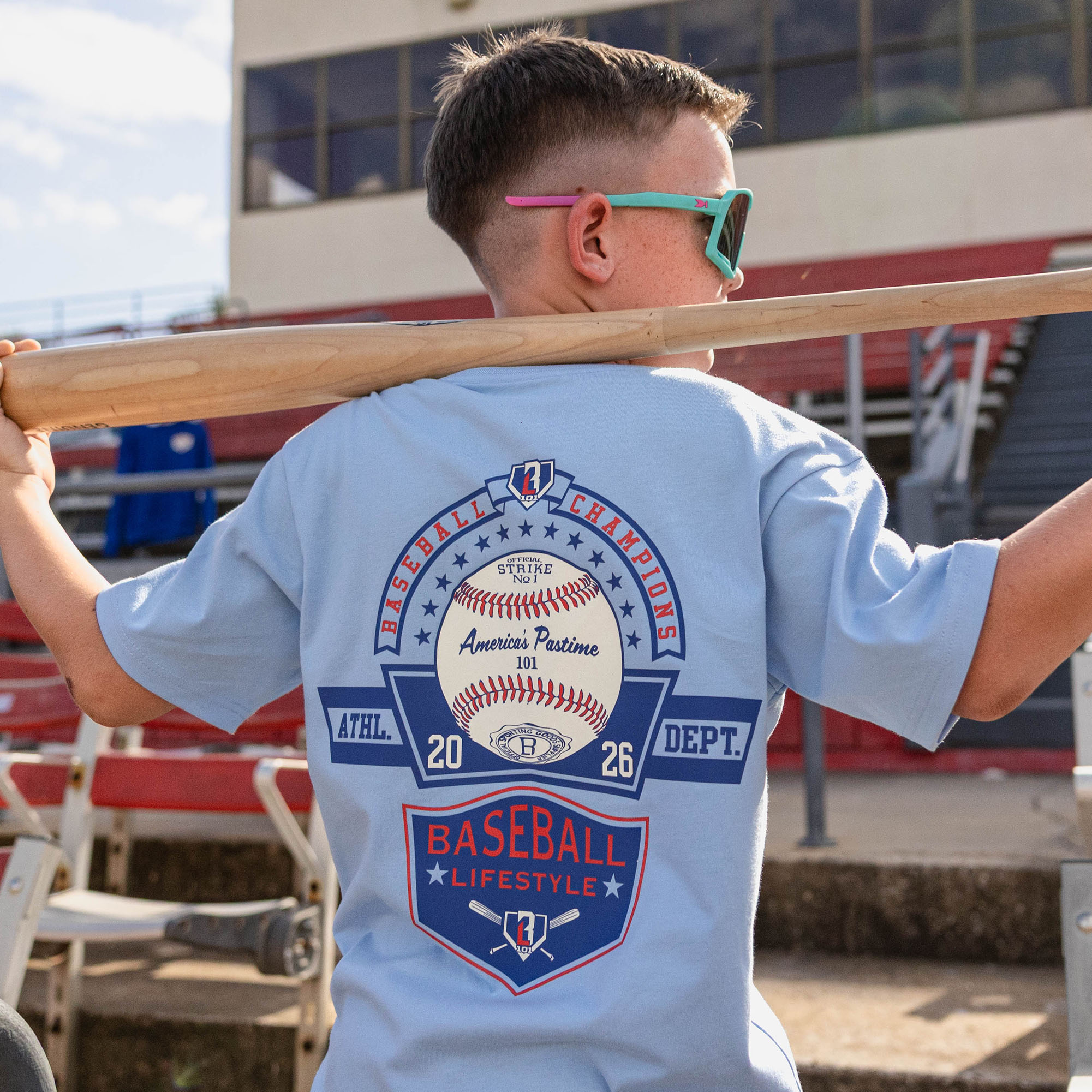 Back view of boy holding a bat across his shoulders at red stadium bleachers wearing a light blue tee with a Baseball Champions America's Pastime 101 back graphic and teal sunglasses