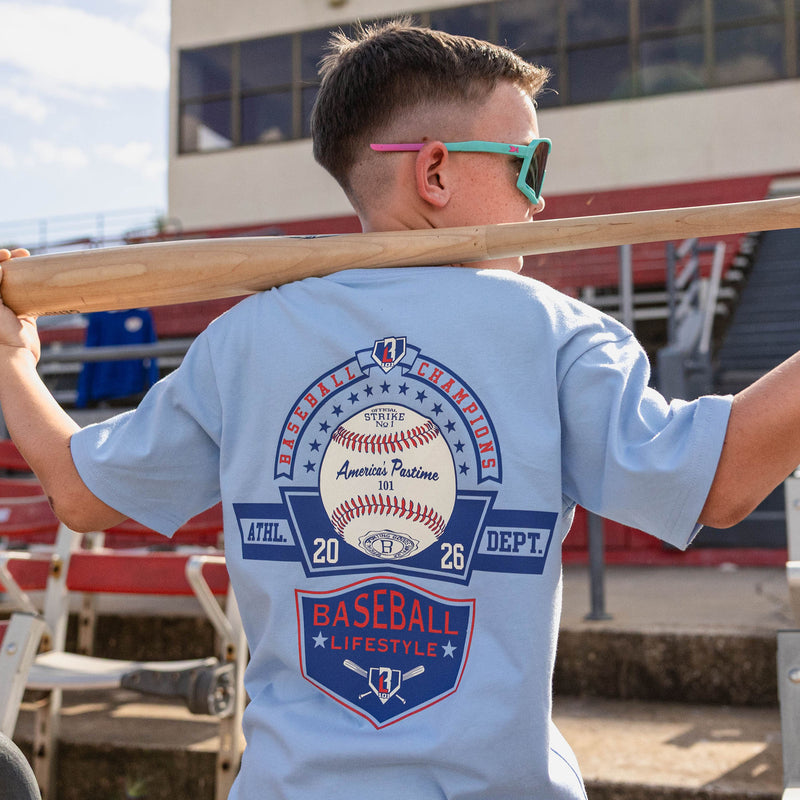 Back view of boy holding a bat across his shoulders at red stadium bleachers wearing a light blue tee with a Baseball Champions America's Pastime 101 back graphic and teal sunglasses