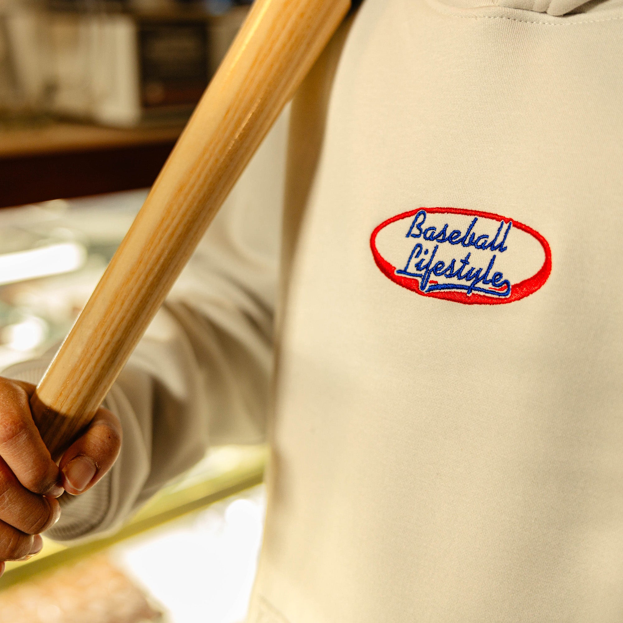 Beige hoodie with 'Baseball Lifestyle' logo held by a person with a baseball bat.