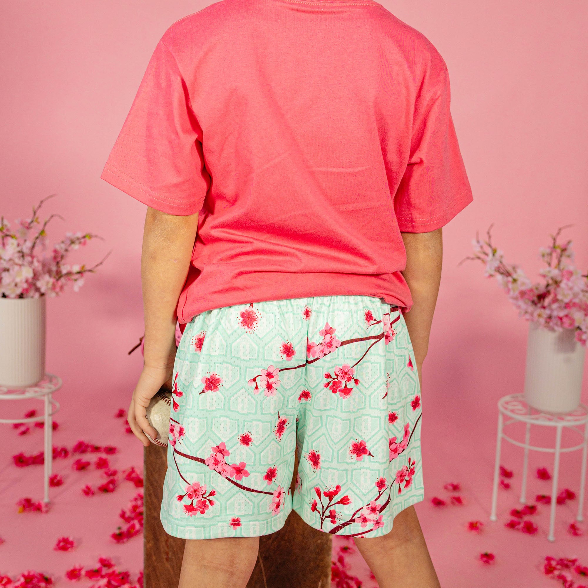 Young boy showing the back of a pink BL101 tee paired with mint green BL101 cherry blossom mesh shorts while holding a baseball against a pink background