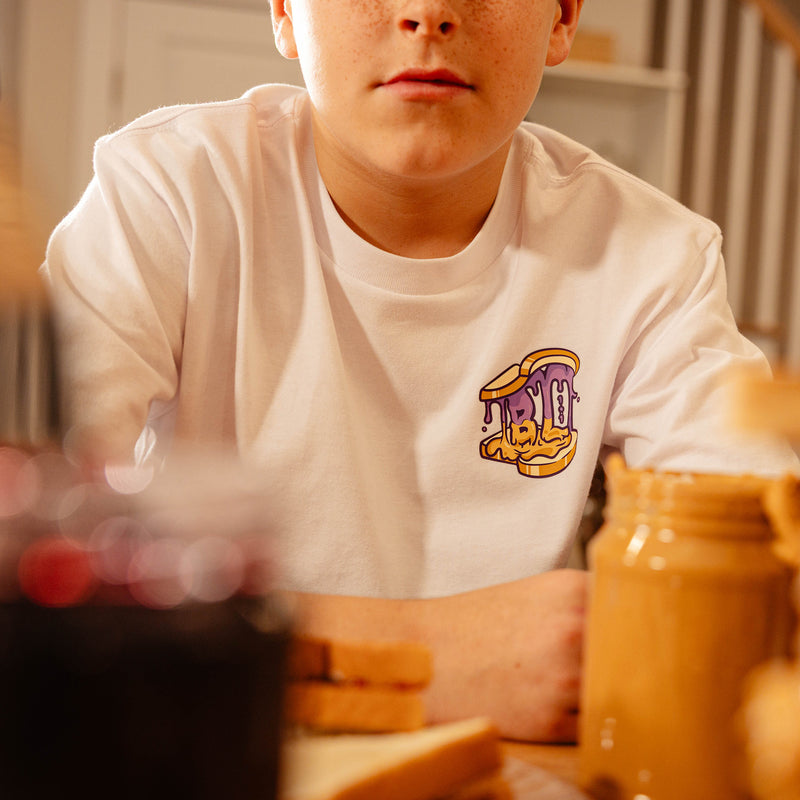 Close-up of the BL101 Peanut Butter and Jelly chest logo on the youth white tee, with a peanut butter jar in the foreground.