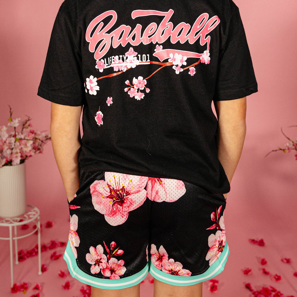 Young boy showing the back of a black BL101 cherry blossom tee paired with black BL101 cherry blossom mesh shorts with teal hem against a pink background