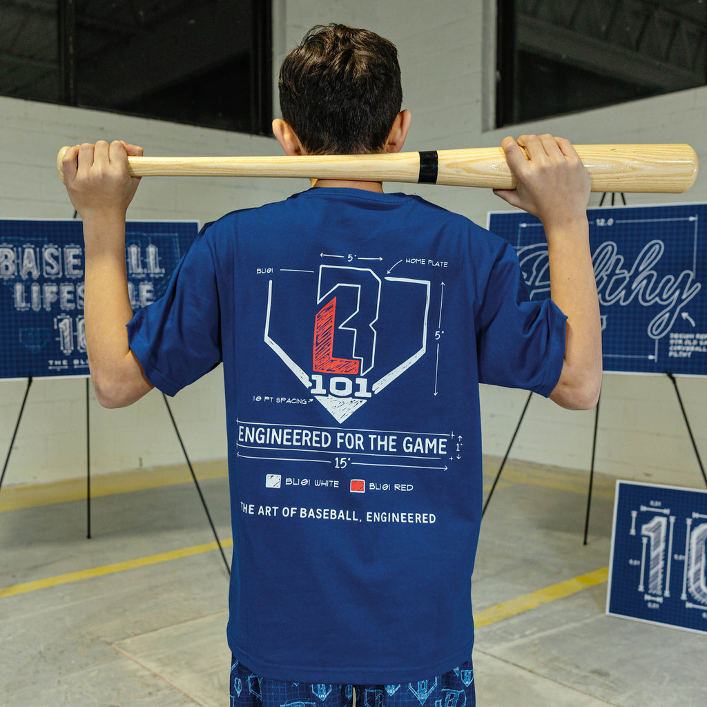 Person wearing a blue t-shirt with a baseball design, holding a bat over their shoulders in an indoor setting.
