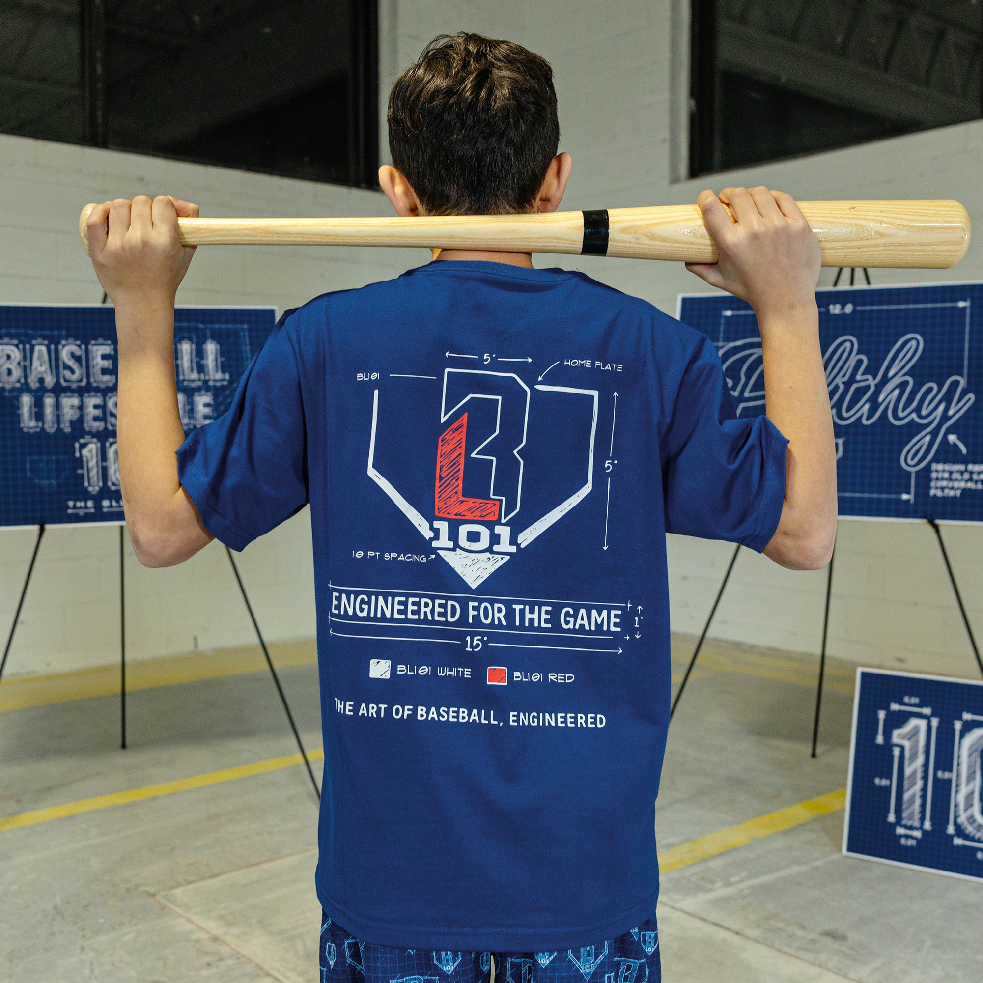 Person wearing a blue t-shirt with a baseball design, holding a bat over their shoulders in an indoor setting.