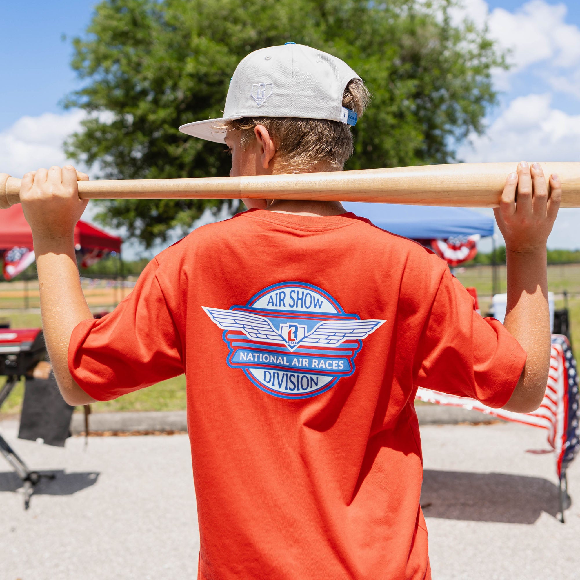Back view of youth male model holding baseball bat across shoulders wearing gray snapback and orange tee with Air Show National Air Races Division graphic on back outdoors