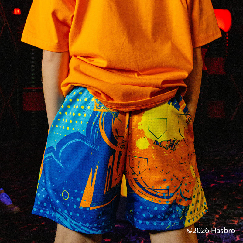 Person wearing bright orange shirt and blue shorts with colorful patterns, standing against a dark background.