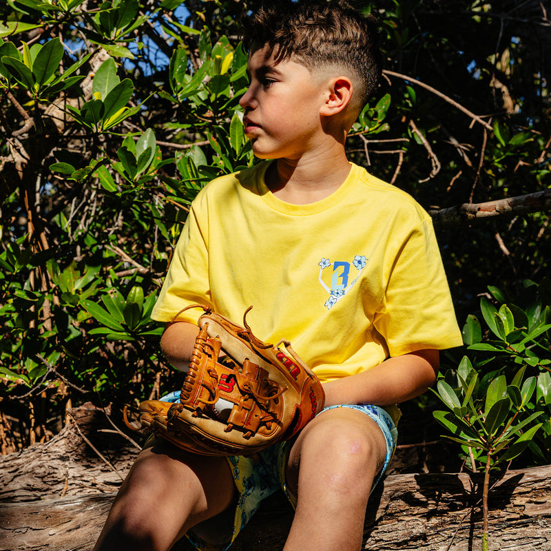 Child in a yellow floral BL101 shirt holding a baseball glove in a natural setting