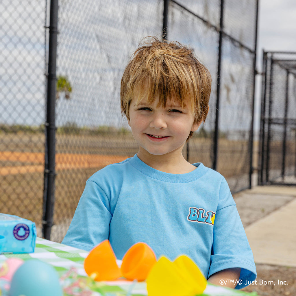 Child in a blue shirt with a peeps BL101 logo, standing outdoors near a table with colorful items.