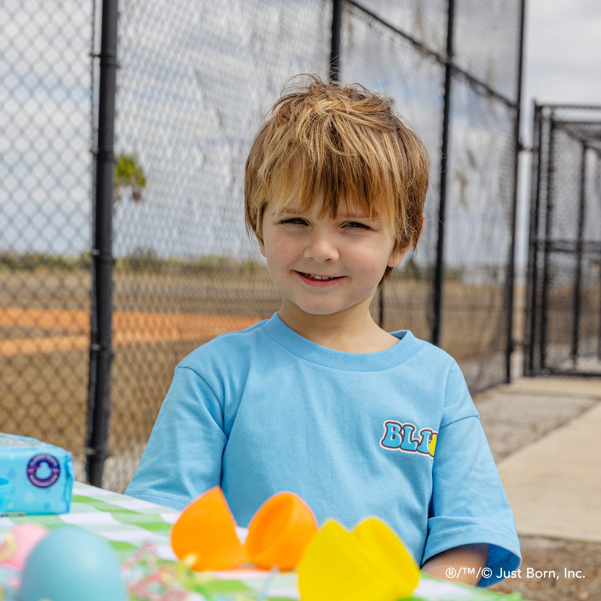Child in a blue shirt with a peeps BL101 logo, standing outdoors near a table with colorful items.