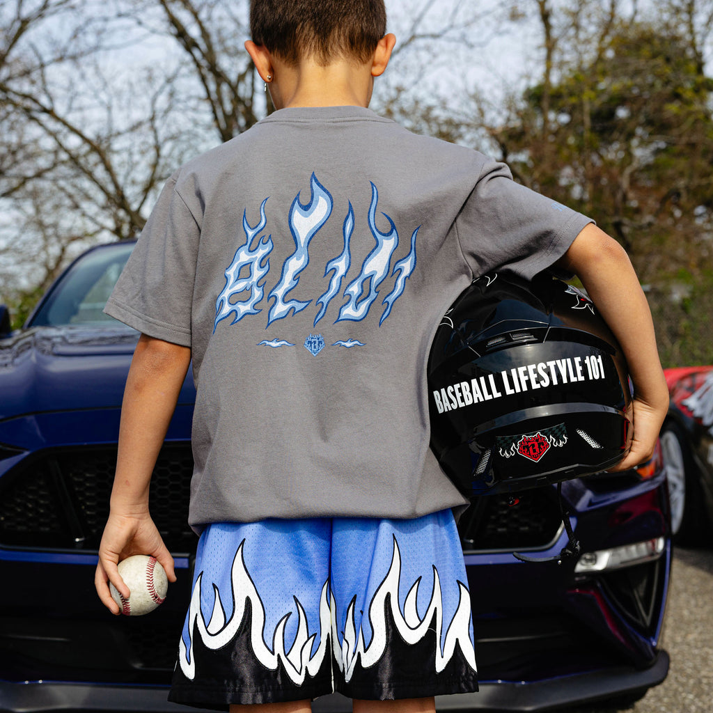 Back view of gray Flame Thrower tee and blue youth flame mesh shorts, boy holding baseball and Baseball Lifestyle 101 helmet beside navy Ford Mustang.
