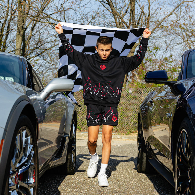 Young male model running between two parked cars while holding a checkered flag overhead wearing a black hoodie with flame stitching and matching black shorts