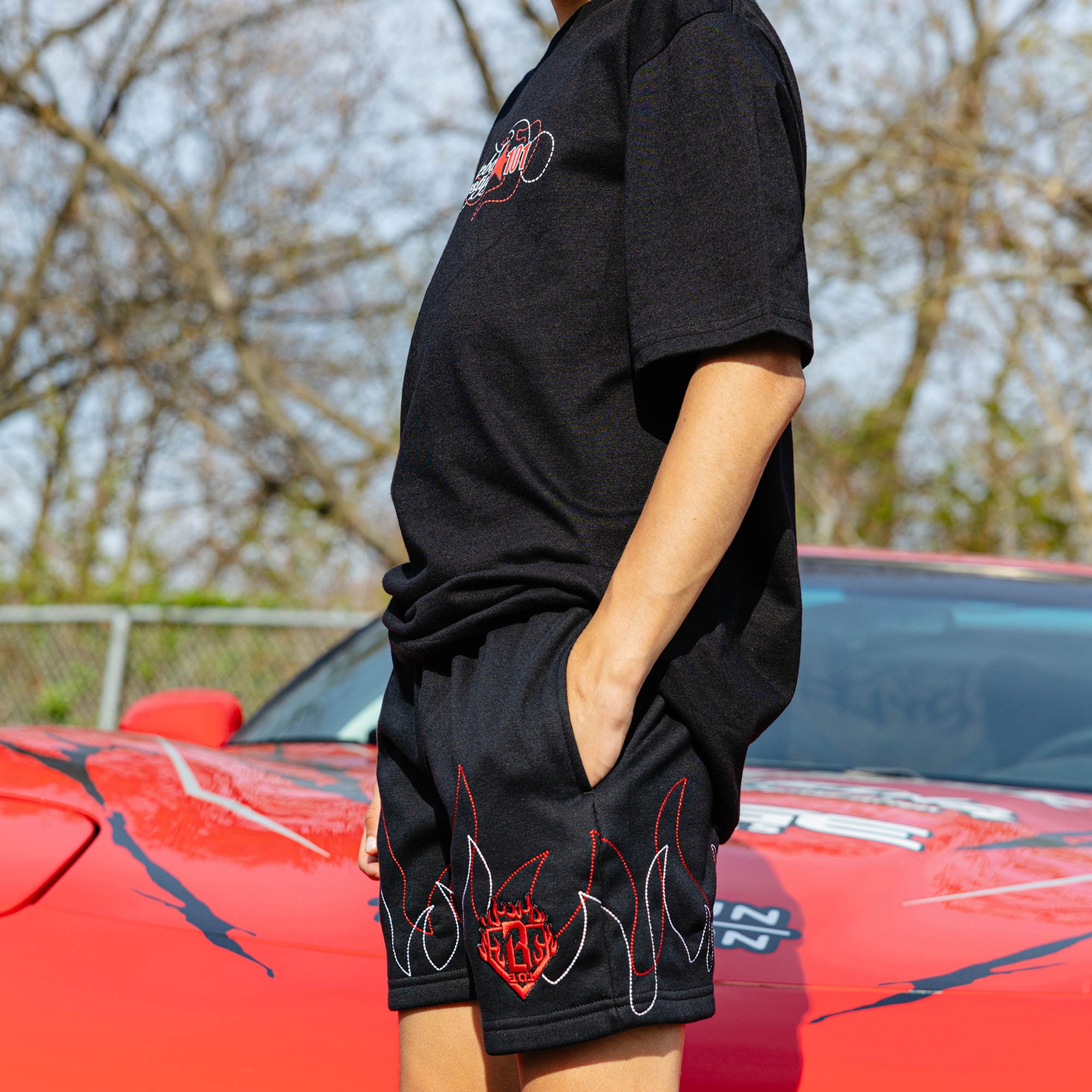 Side view of young boy in black Flames Stitched tee and sweatshorts with hand in pocket, standing beside red car.