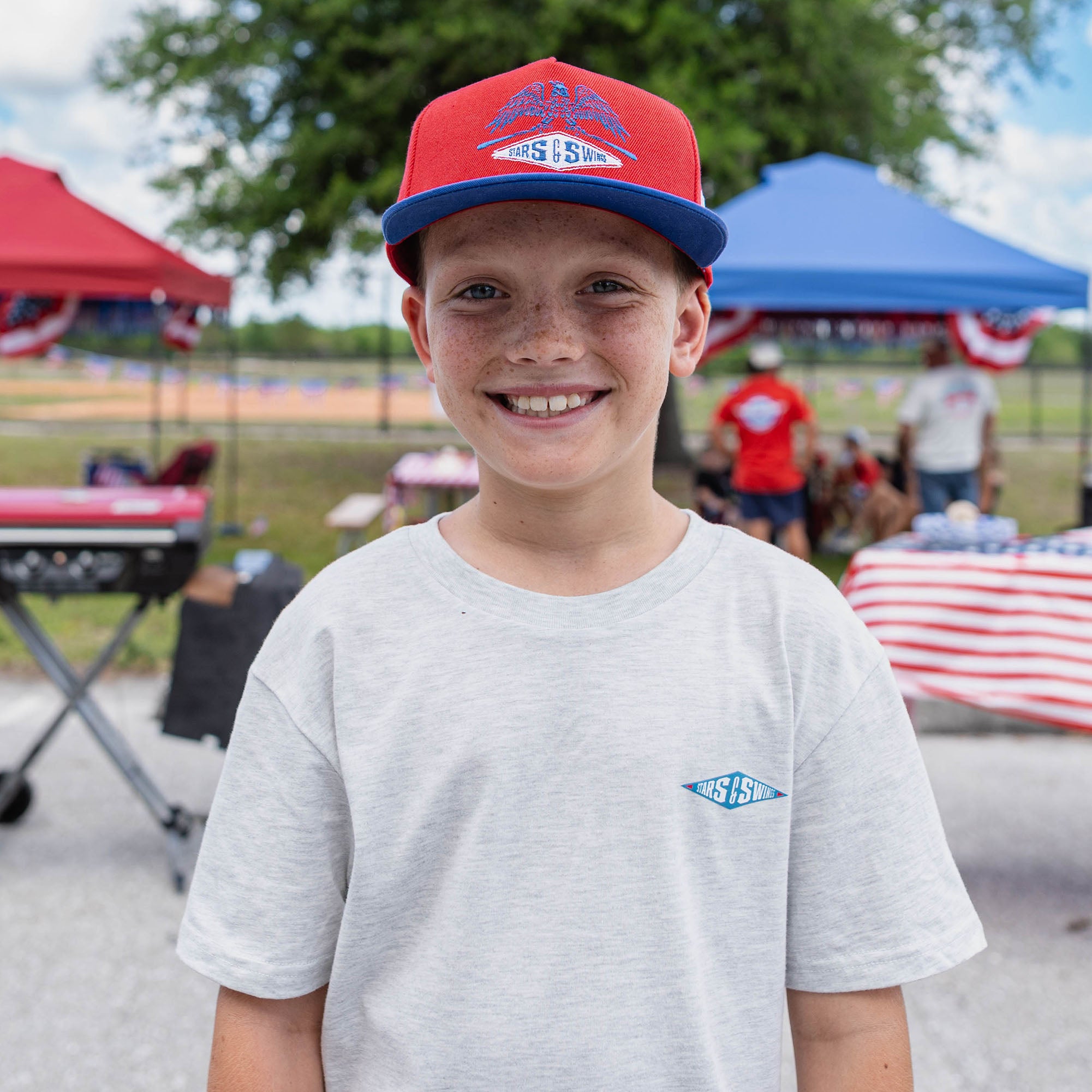 Young boy wearing a red and blue cap with a logo, standing outdoors with tents and people in the background.