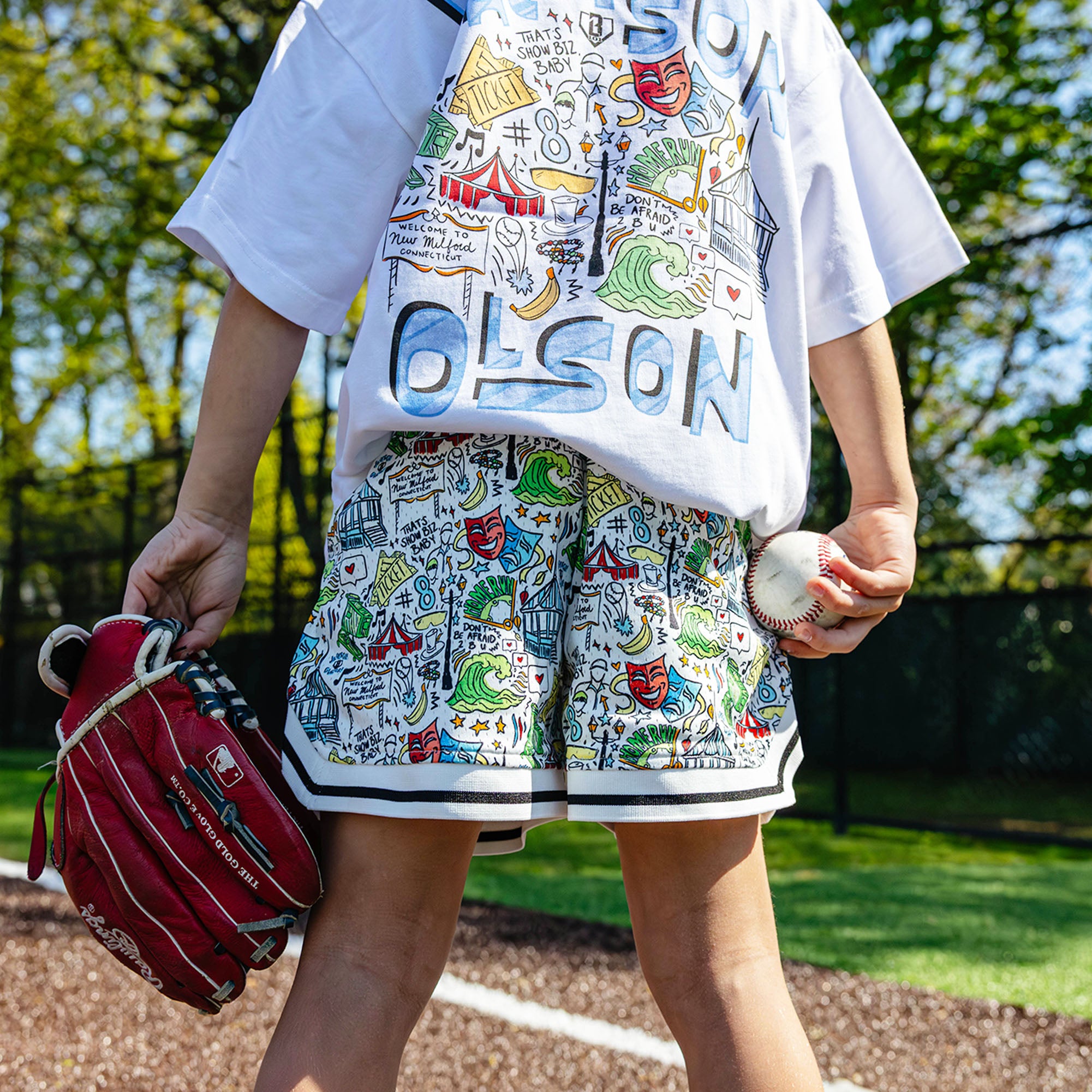 Youth player holding a ball and glove on a baseball field, showing the white Jackson Olson x BL101 Hometown Shorts paired with the Hometown Relaxed Tee back graphic