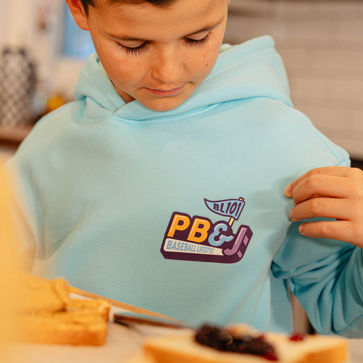 Close-up of the PB&J Baseball Lifestyle chest logo on the youth light blue hoodie, with a young boy making a sandwich in the foreground.