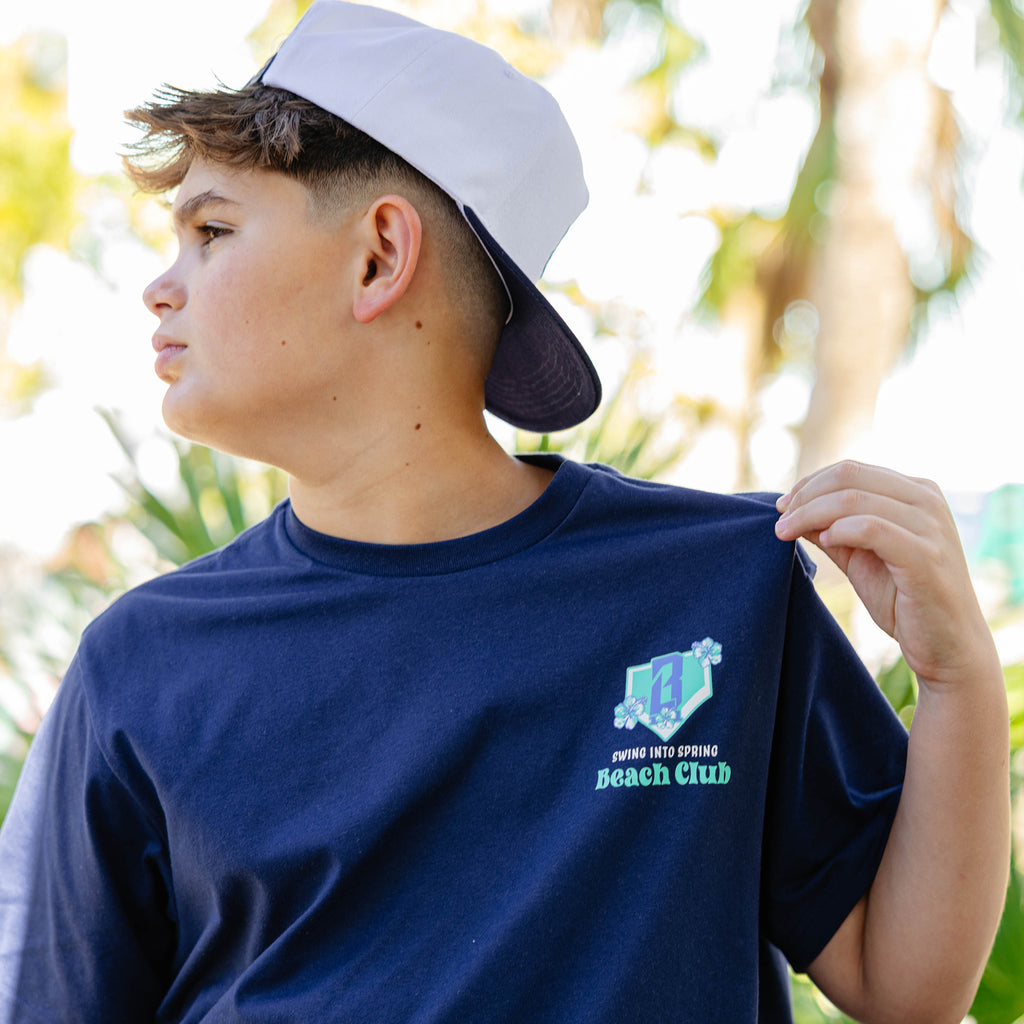 Person wearing a navy blue t-shirt with a floral BL101 logo and 'Swing into Spring Beach Club', standing outdoors with blurred greenery in the background