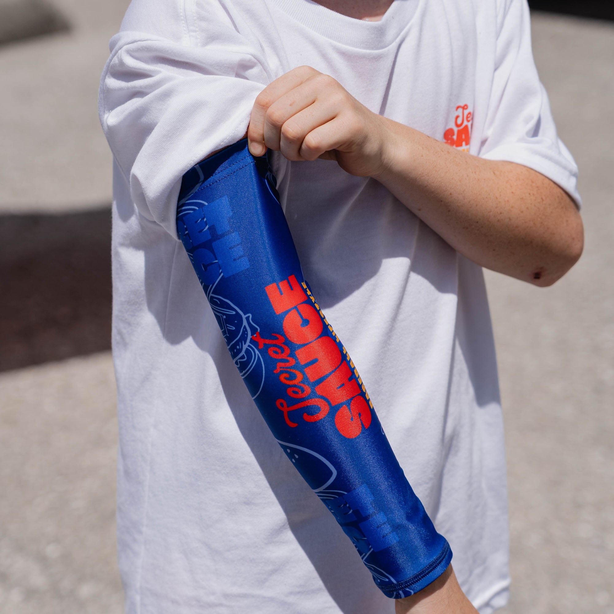 Youth model holding blue Secret Sauce arm sleeve extended showing full Secret Sauce logo and burger line art print while wearing white Secret Sauce tee outdoors