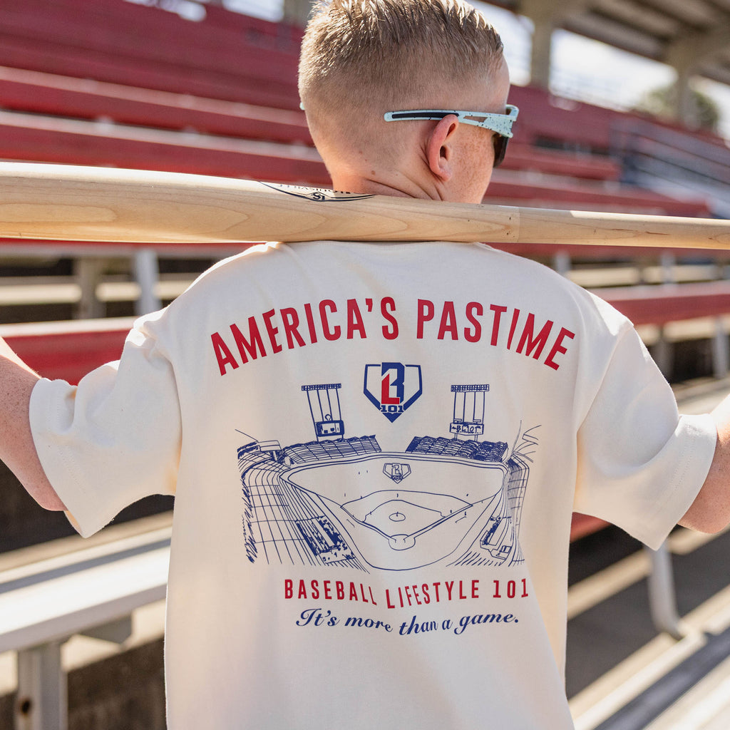 Back view of the BL101 youth America's Pastime cream tee on a young boy in a stadium holding a baseball bat, showing the stadium sketch graphic and "It's more than a game" text.