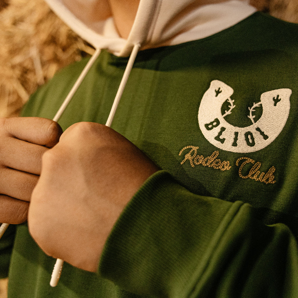 Green hoodie with horseshoe embroidered logo and 'BL101 Rodeo Club' text, held by a person.