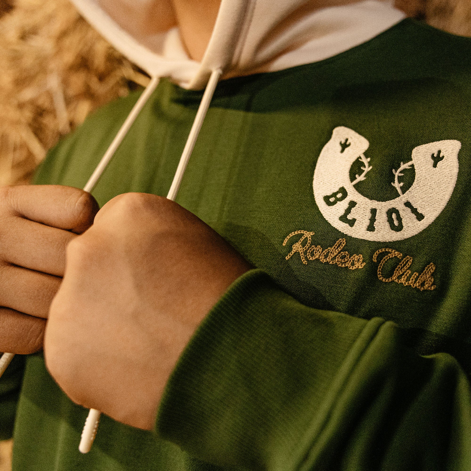 Green hoodie with horseshoe embroidered logo and 'BL101 Rodeo Club' text, held by a person.
