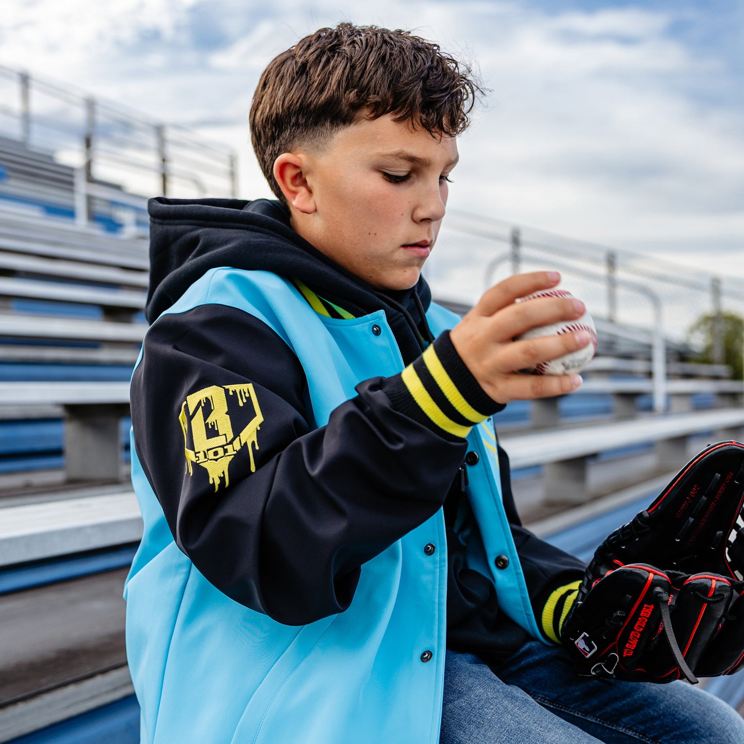 Young boy in a blue, yellow and black jacket with a yellow BL101 logo on the sleeve sitting on bleachers holding a baseball and glove.