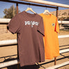 Two t-shirts, one brown with cow print and one orange,  hanging on a metal railing with a desert landscape in the background.