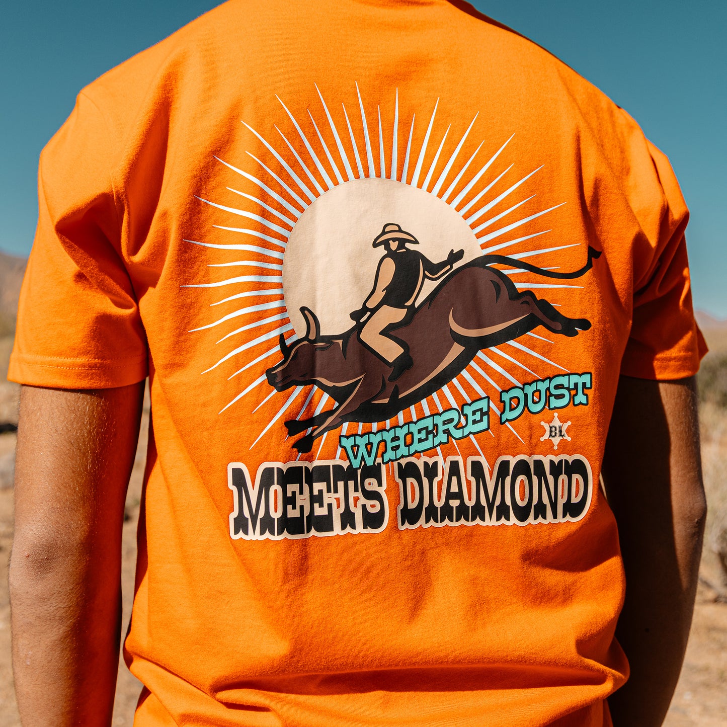 Orange t-shirt with a graphic design of a person riding a bull and text 'Where Dust Meets Diamond' in a desert setting.