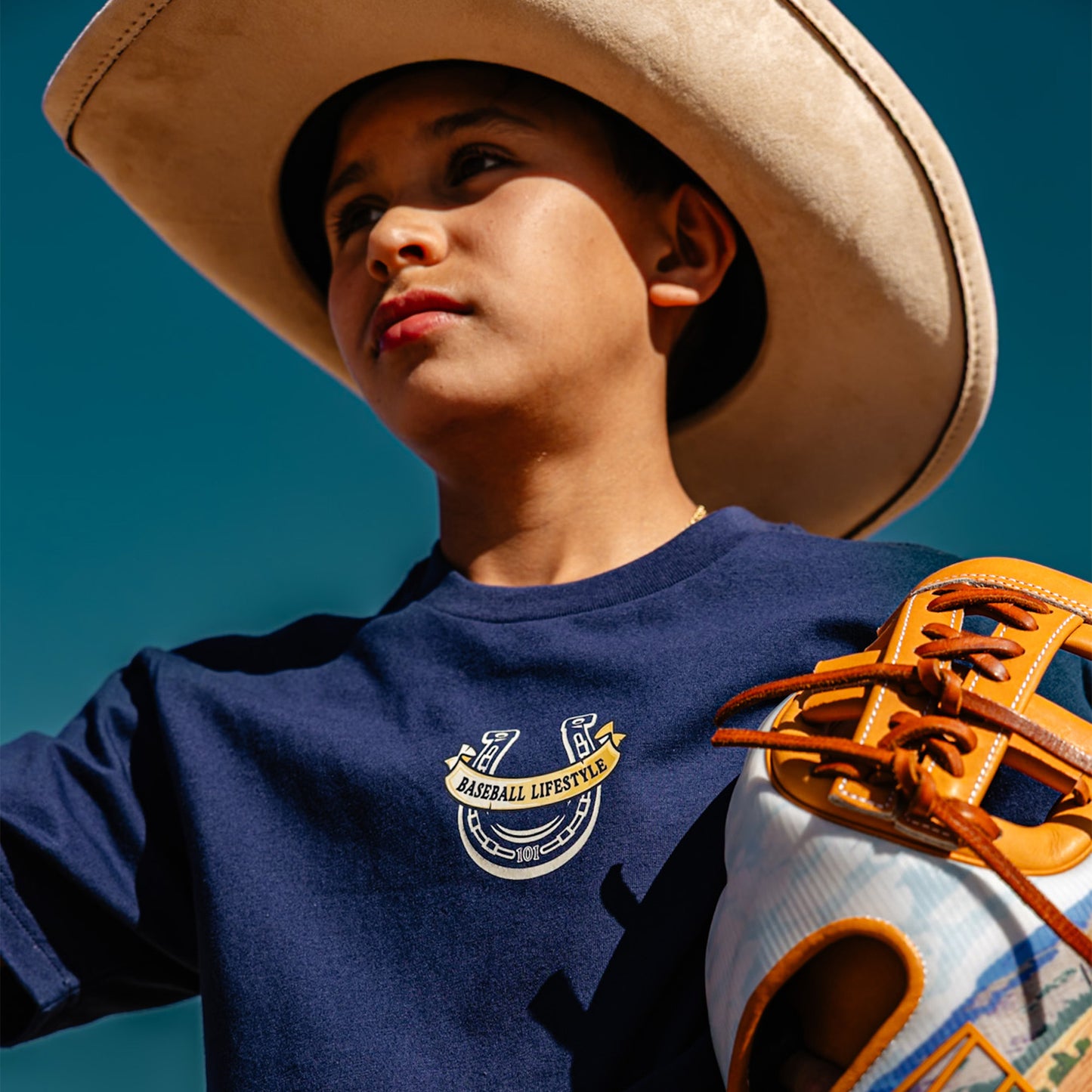Child wearing a blue t-shirt with a Baseball Lifestyle horseshoe logo and a baseball glove, against a clear blue sky.
