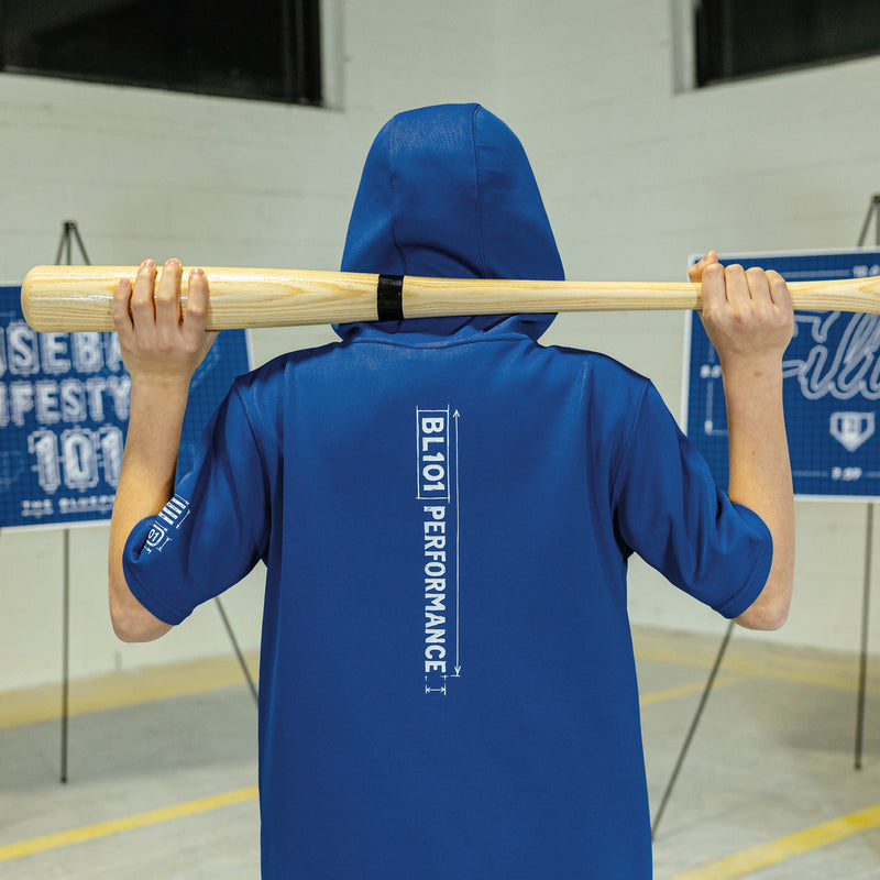 Person wearing a blue hooded jacket with 'BL101 Performance' branding, holding a baseball bat indoors.