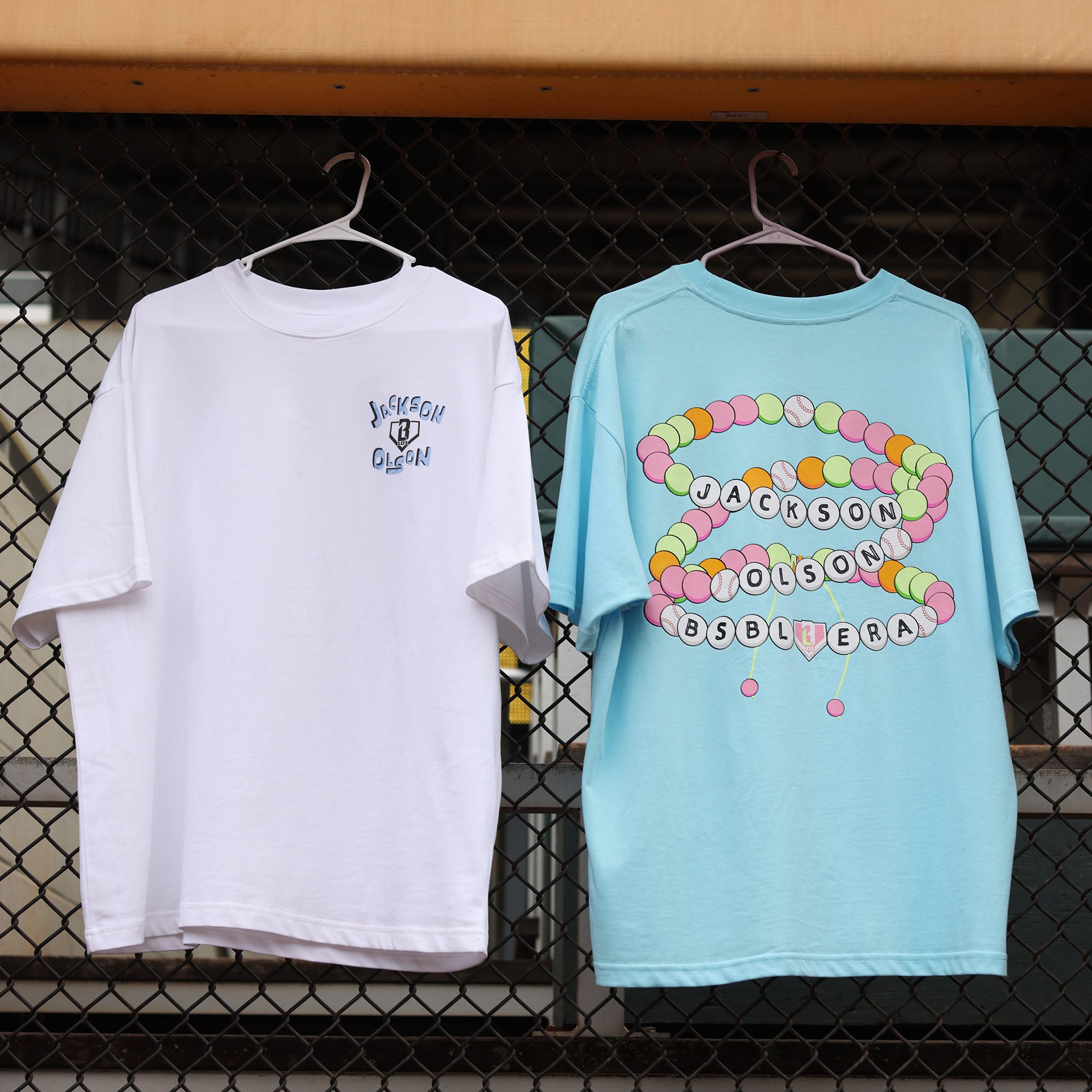 White and light blue Jackson Olson x BL101 youth relaxed tees hanging on a chain-link fence, showing front and back graphic details side by side