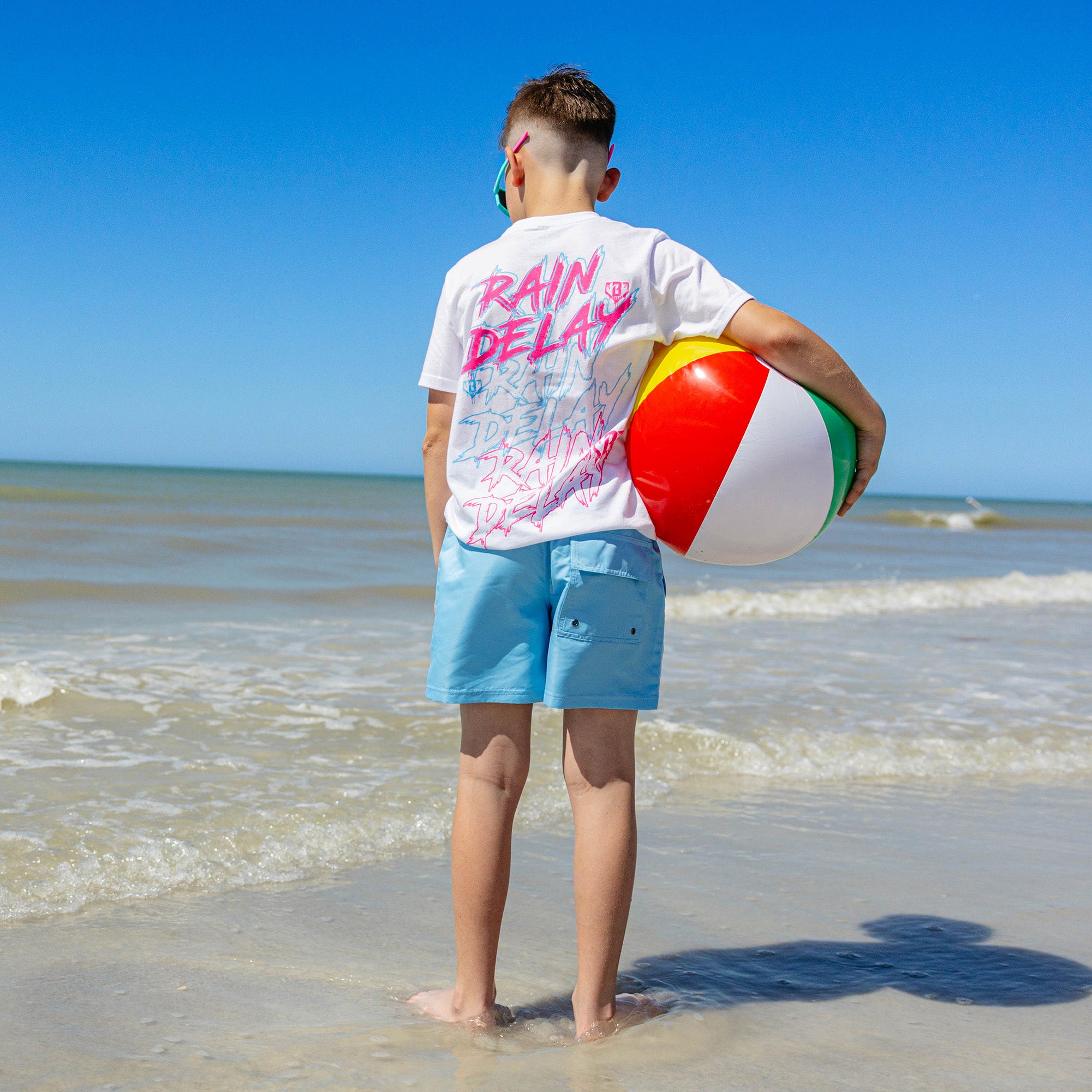 Back view of a young boy in light blue BL101 youth swim trunks and white Rain Delay graphic tee, standing at the water's edge holding a beach ball.